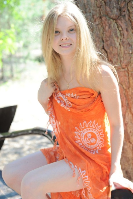 Young blonde Kisa removes her wrap to pose artistic on bench under an old tree